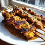 Drie gegrilde spiesjes met malse Chicken Satay, overgoten met pindasaus, op een wit bord.