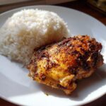 Close-up van een sappige, gekruide kipdijfilet naast een berg witte rijst op een wit bord.