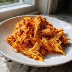 Een close-up van draadjesvlees van Slow Cooker Buffalo Chicken, bedekt met een oranje saus, geserveerd op een wit bord.