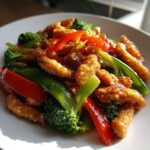 Geglaceerde stukjes kip en kleurrijke groenten zoals broccoli, rode paprika en peultjes in een Chicken Stir Fry op een witte schotel.