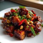 Close-up van glanzende, gekarameliseerde blokjes Kung Pao Tofu op een witte schotel, gegarneerd met pinda's en lente-ui.