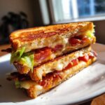 Een doormidden gesneden BLT Egg Grilled Cheese Sandwich met gesmolten kaas, tomaat, sla en spek op een wit bord.