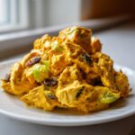 Close-up van een portie romige, gele Curry Chicken Salad met rozijnen en selderij op een wit bord.