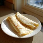 Twee opgerolde, lichtbruin gebakken Egg White Wraps liggen op een wit bord bij het raam.