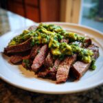 Plakjes gegrilde flank steak royaal overgoten met heldergroene avocado chimichurri saus op een wit bord.