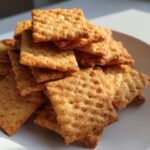 Close-up van een stapel goudbruine, vierkante Homemade Cheese Crackers op een wit bord.