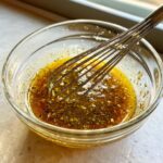 Close-up van een glazen kom met olieachtige, gekruide vloeistof die wordt gemengd met een garde voor de Homemade Italian Salad Dressing.