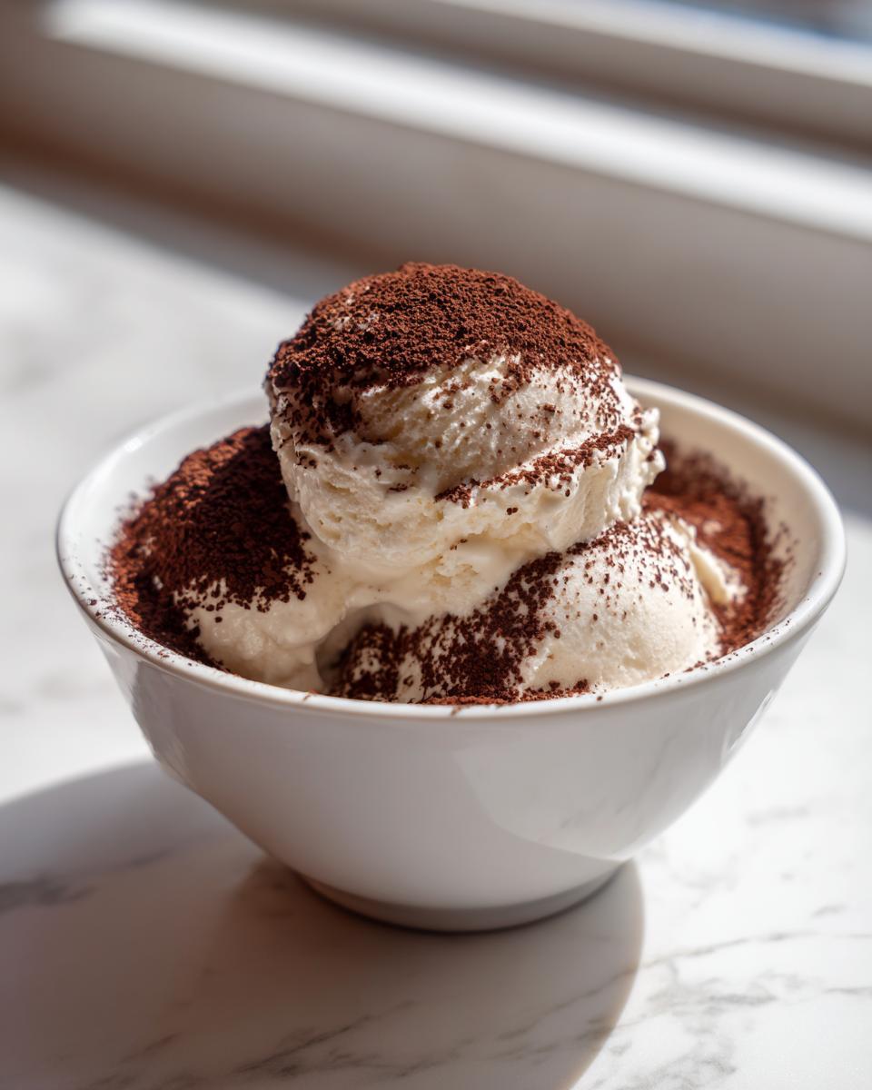 Drie bollen romige Tiramisu Ice Cream in een witte kom, rijkelijk bestrooid met cacaopoeder.