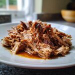 Close-up van sappig, uit elkaar getrokken Slow Cooker Pulled Pork met jus op een wit bord.