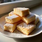 Stukjes luchtige Shortbread Cookies gestapeld op een wit bord, bestrooid met poedersuiker.