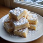 Een stapel vierkante Shortbread Cookies rijkelijk bestrooid met poedersuiker op een wit bord.