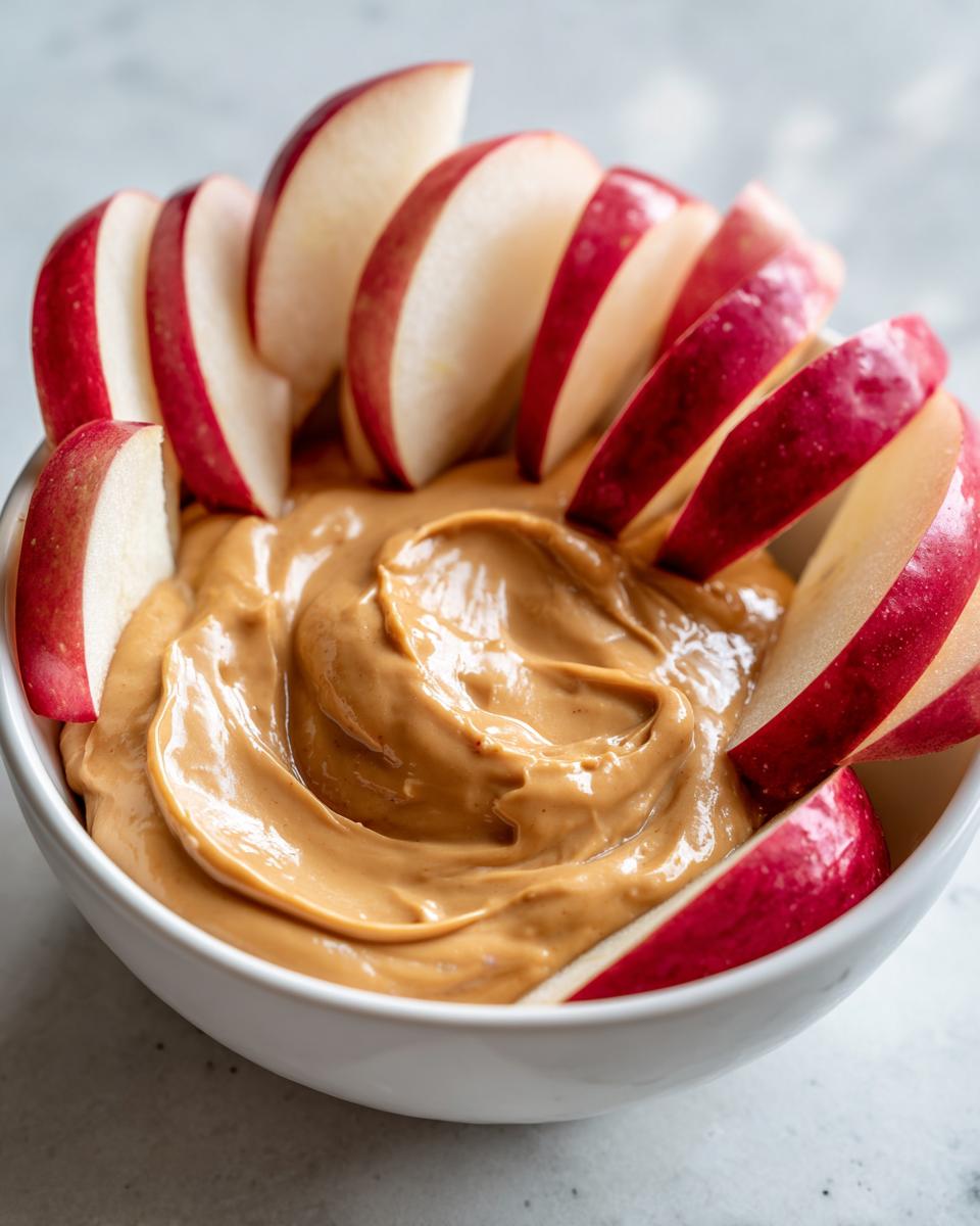 Een kom met romige Toffee Apple Dip, omringd door verse, rode appelpartjes.
