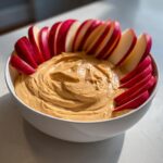 Een witte kom gevuld met romige Toffee Apple Dip, omringd door rode, gesneden appels.