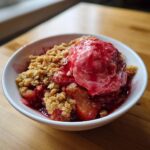 Een kom met warme Strawberry Rhubarb Crumble, getopt met een bol roze ijs.