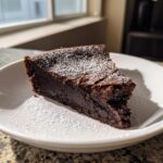 Een heerlijke, donkere punt Tunnel Of Fudge Cake bestrooid met poedersuiker op een wit bord.