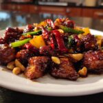 Close-up van een bord met glanzende, gekarameliseerde Kung Pao Chicken stukjes, pinda's, rode pepers en groenten.