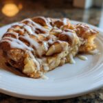 A close-up of a slice of Apple Cinnamon Roll Bake drizzled generously with white icing on a white plate.