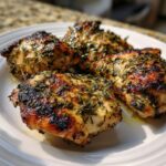 Four pieces of perfectly grilled Greek Lemon Chicken thighs generously coated in herbs on a white plate.