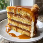 A tall slice of Salted Caramel Butterscotch Cake showing multiple layers of sponge and filling, generously topped with dripping caramel sauce.