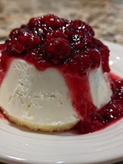 A creamy white Panna Cotta With Berry Sauce served on a white plate, topped with rich red berry compote.