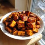 A white plate piled high with perfectly roasted, seasoned Sweet Potato Bites, illuminated by sunlight near a window.