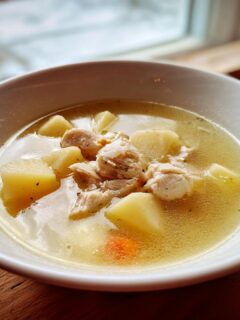 A close-up of a white bowl filled with clear broth, chunks of chicken, and large pieces of potato in Chicken Soup With Potatoes.