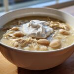 A close-up of a white bowl filled with creamy White Chicken Chili, topped with a dollop of sour cream.