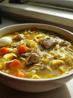 A close-up of a white bowl filled with rich, yellow Chicken Noodle Soup featuring chunks of chicken, wide egg noodles, carrots, and celery.
