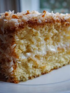 A close-up of a moist slice of Hawaiian Dream Cake, layered with cream and topped with toasted coconut flakes.