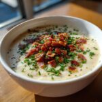 A close-up of a bowl of creamy Outback Potato Soup garnished generously with crispy bacon bits and fresh chopped chives.