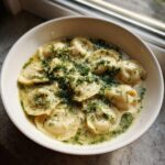 A close-up of a white bowl filled with Pierogi Dill Soup, featuring dumplings in a creamy broth topped generously with fresh dill.